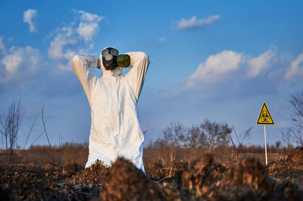 Male environmentalist clutching his head in despair while sitting at damaged grass and soil after fire. Scientist in gas mask looking at the sky at burnt field. Concept of ecology and burned earth. - Photo, Image