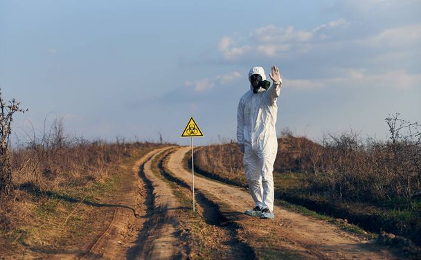Man in white coverall, gas mask, gloves standing on road in field next to caution symbol showing stop sign, on a sunny day. Biohazard sign warning about harmful biological substances and danger - Photo, Image