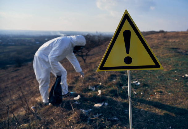 Close up of yellow triangle with black exclamation mark warning about danger and hazards. Warning sign with ecologist in radiation suit picking up garbage on blurred background. Concept of ecology - Photo, Image