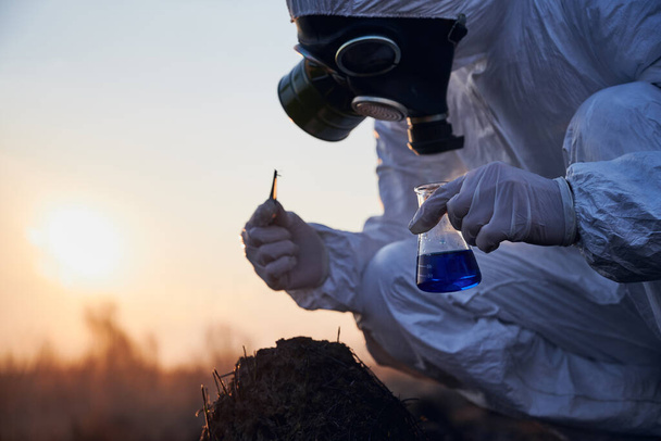 Zijaanzicht, close-up momentopname van een gehurkte ecoloog in een wit pak, gasmasker en handschoenen die monsters verzamelt van verschroeide mierenhoop, met glazen kolf met blauwe vloeistof en pincet bij zonsondergang - Foto, afbeelding