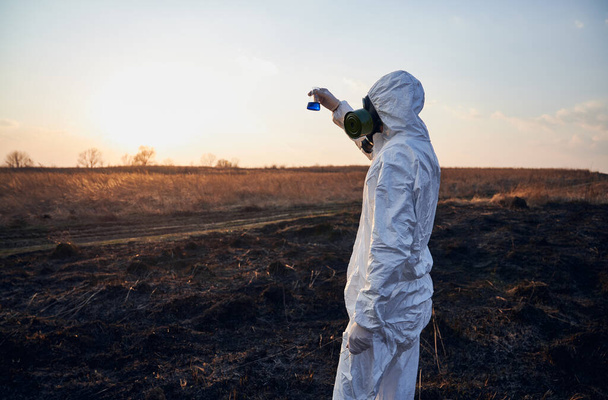 Laboratorní pracovník v bílé kombinéze, plynová maska a rukavice pozorovající zkumavku s modrou tekutinou při studiu spálené trávy a půdy na spáleném území na zapadajícím slunci, koncepce ekologické katastrofy - Fotografie, Obrázek