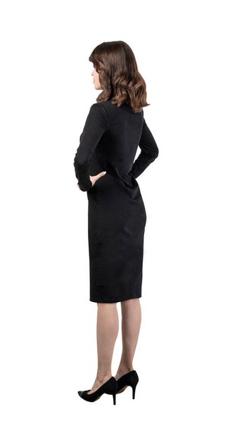 Isolated full length portrait of serious confident young European businesswoman with long wavy dark hair wearing black dress and standing with hands on her waist - Fotó, kép