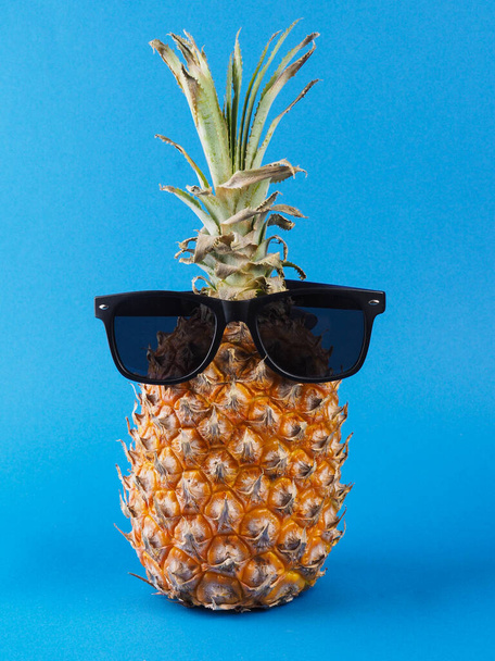 Front view of a ripe orange pineapple with green leaves and black sunglasses on it, on a blue background. - Fotó, kép
