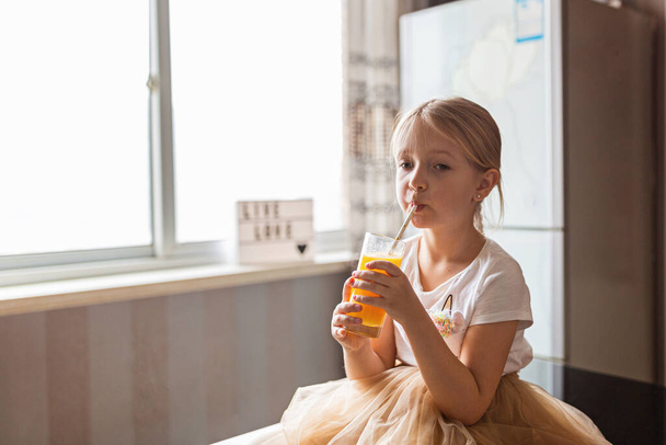 Cute little caucasian girl with blonde hair sitting on the kitchen table and drinking fresh mango juice. Healthy lifestyle concept. Kid 6 years old - Fotó, kép