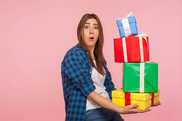 Portrait of funny girl carrying lot of heavy present boxes and looking at camera with amazed comical expression, surprised by many birthday gifts. indoor studio shot isolated on pink background - 写真・画像