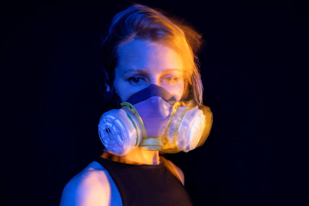 Beautiful girl in a respirator looking down. air pollution and virus theme. expressive beautiful eyes look. Dark background. Emotional portrait about pandemic and disaster survival. Yellow and blue - Фото, изображение