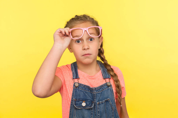 Portrait of amazed stylish little girl in denim overalls taking off pink eyeglasses and looking with astonished expression, child surprised by her vision improvement. indoor studio shot, isolated - Photo, image