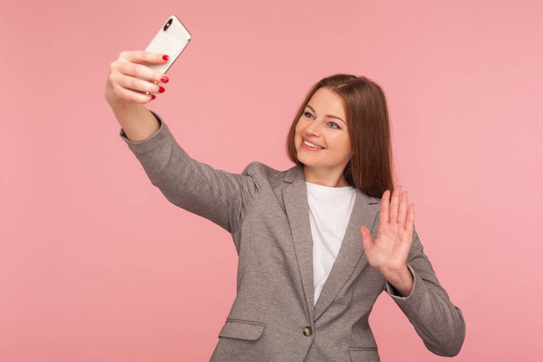 Portrait of charming businesswoman in suit jacket making video call with hello gesture, having online conference using cellphone, corporate cellular and mobile communication. studio shot isolated - Fotoğraf, Görsel