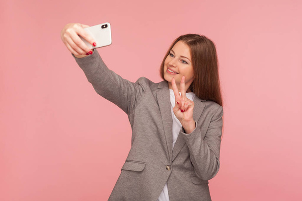 Portrait of successful businesswoman in suit jacket making video call with victory gesture, having online conference using cellphone, corporate cellular and mobile communication. studio shot - Zdjęcie, obraz