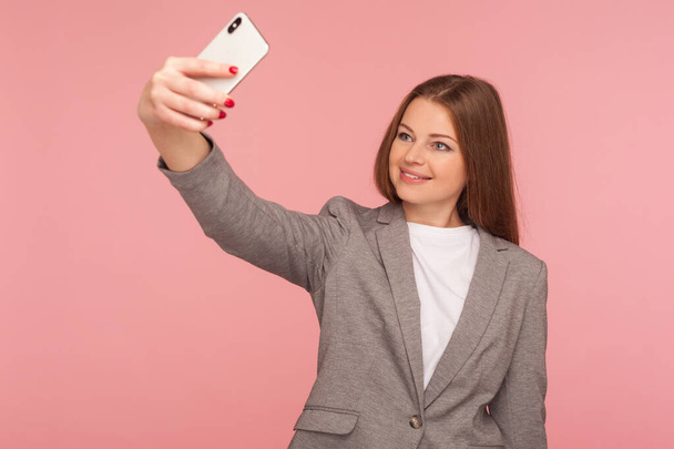 Portrait of attractive businesswoman in suit jacket making video call, having online conference using cellphone, corporate cellular and mobile communication. studio shot isolated on pink background - Zdjęcie, obraz