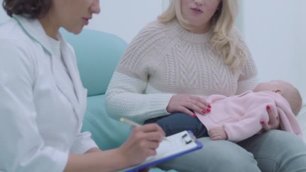 Female pediatrician writing information, talking to mother of the baby, health - Кадры, видео