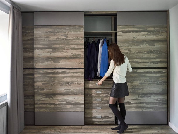 Stylish young woman standing with her back looks into an open wardrobe with clothes and opens drawer. Choosing an outfit at home. - Photo, Image