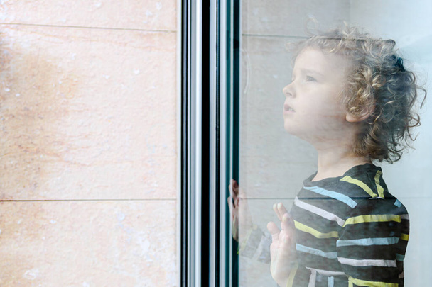 ξανθιά κοριτσάκι κοιτάζει έξω από το παράθυρο κατά τη διάρκεια του εγκλεισμού COVID-19 - Φωτογραφία, εικόνα