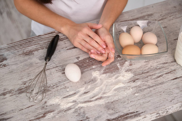 нерозпізнана жінка готує в білому одязі готує на кухні. На столі миска з яйцями, пляшка молока і віник. текстура дерева
 - Фото, зображення