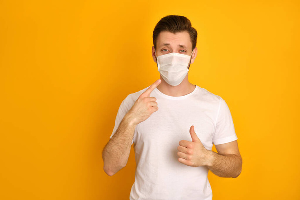 Caucasian young guy wears face white mask to protect himself against covid19, wants to be healthy, has a white t-shirt on, points with his finger at the mask - Foto, imagen