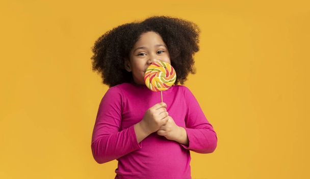 Positive little afro girl licking lollipop candy, enjoying sugar treat - Foto, imagen