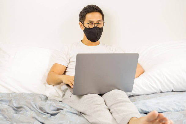 Mies, jolla on suojaava naamio, työskentelee kannettavalla tietokoneella sängyssä kotona covid-19-epidemia
 - Valokuva, kuva