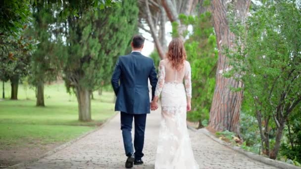 Back view of amazing just married couple walking happily in the park. Handsome groom in dark blue suit and pretty red hair bride in white lace dress holding hands together and walking in slow motion - Footage, Video