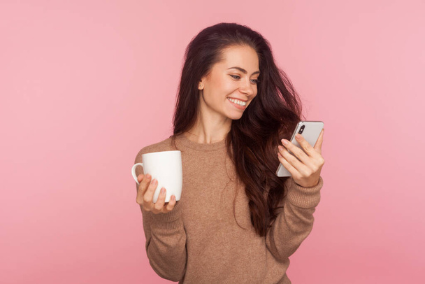 Portrait of stylish attractive young woman with long brown hair holding cup of coffee and watching message on smartphone, having break and relaxing. indoor studio shot isolated on pink background - Valokuva, kuva