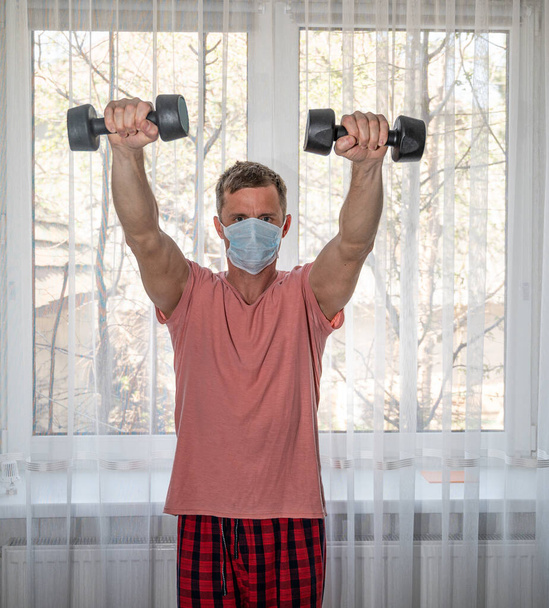 Quarantine time concept. Active man in protective mask exercising with dumbbells at home - Fotografie, Obrázek