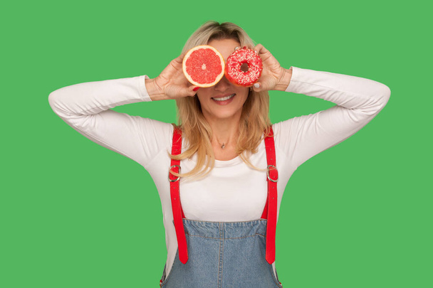 Healthy fruits vs junk food. Portrait of playful stylish woman covering eyes with doughnut and fresh juicy grapefruit, having fun, making choice. indoor studio shot isolated on green background - Zdjęcie, obraz