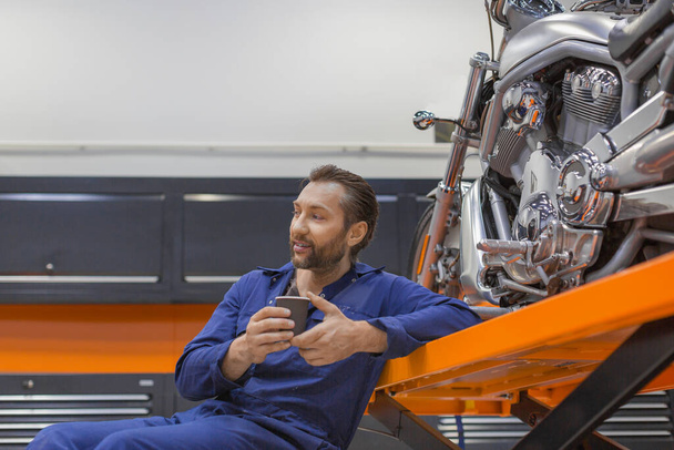 Un hombre sonriente en el taller se sienta cerca de una motocicleta y bebe café
. - Foto, Imagen