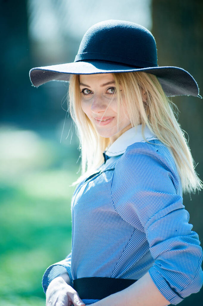 young pretty blond woman in blue dress and black hat posing on park on sunny day background - Фото, зображення
