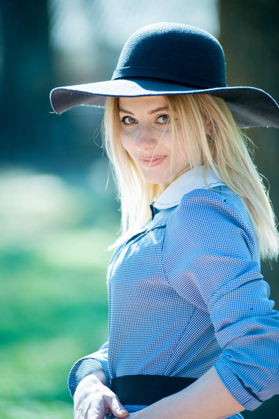 young pretty blond woman in blue dress and black hat posing on park on sunny day background - Photo, Image