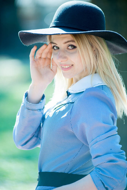 young pretty blond woman in blue dress and black hat posing on park on sunny day background - 写真・画像