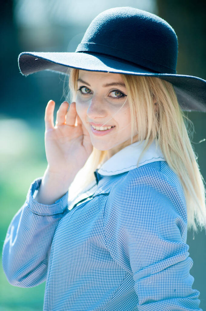Genç, sarışın, mavi elbiseli, siyah şapkalı, güneşli bir günde parkta poz veren bir kadın. - Fotoğraf, Görsel
