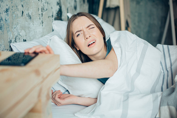 Young woman lying in bed and opening one eye to get her ringing phone in the morning - Photo, Image