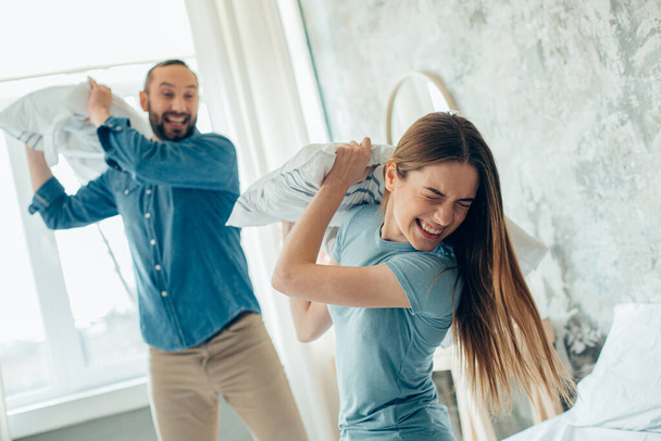 Funny young man and woman laughing and having a real pillow fight on the bed - 写真・画像