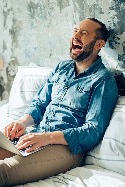 Jeune homme avec un ordinateur portable assis sur le lit et fermant les yeux tout en riant
 - Photo, image