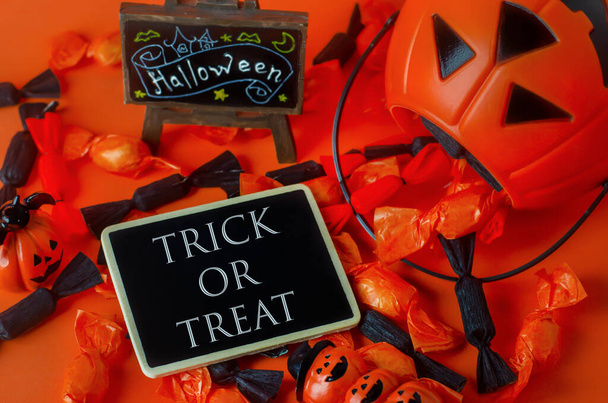 Fondo de Halloween - Truco o trato en etiqueta de madera negra con bolsa de calabaza y caramelos negros y naranjas sobre fondo naranja
 - Foto, Imagen