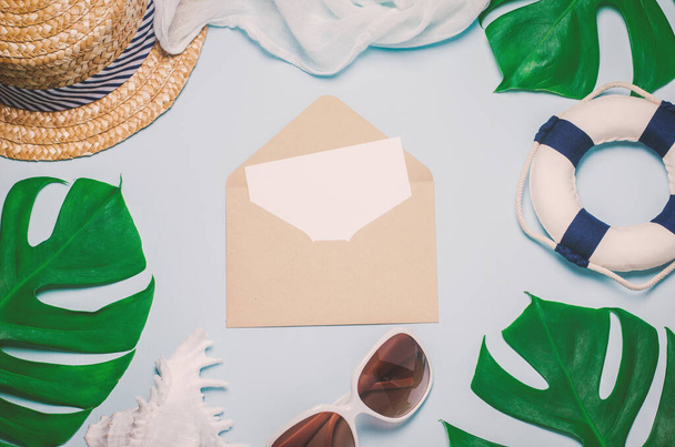 Blank white greeting card and brown envelope with straw hat, Seashell, sunglasses, lifebuoy and green leaves on blue wooden background with vintage tone - Summer background - Fotografie, Obrázek