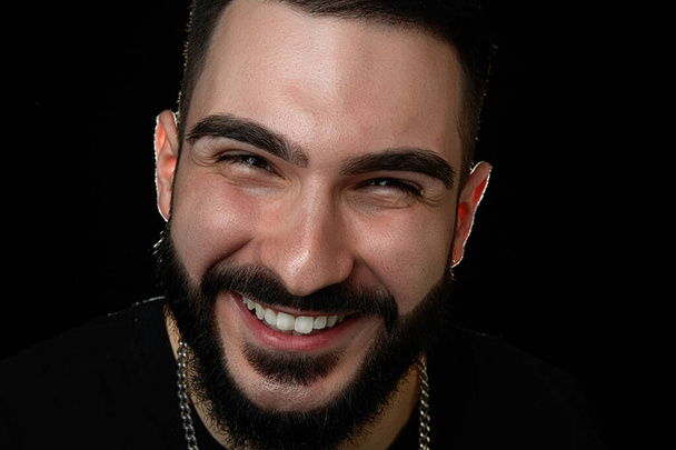 close-up de um retrato dramático de um jovem sorridente, músico, cantor, rapper com barba em roupas pretas. sobre um fundo isolado preto
. - Foto, Imagem
