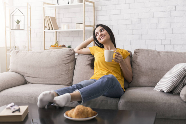 Zostań w domu, napij się kawy. Zrelaksowana Latynoska dziewczyna ciesząca się poranną kawą na kanapie w salonie - Zdjęcie, obraz
