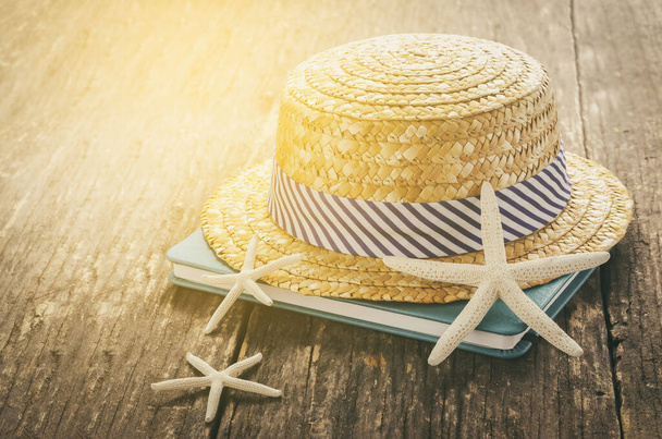Beach concept - Straw Hat with white and blue striped on wooden table with book and starfishes with sunlight and vintage tone - Photo, Image