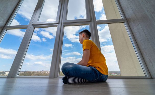 Boy sitting at floor at balcony. Kid looking at nature outside window. isolation at home while quarrantine. Sad boy dreaming. - Foto, Imagen