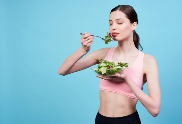 Fitness young cute girl, with brown hair, in a beige sports top, closing her eyes with enjoying eating a vegetarian salad, standing on a blue background. The concept of sports and diet, weight loss. - Photo, Image