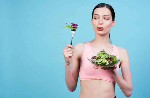 Young brown-haired athletic cute girl with blue eyes, in a beige sports top, holds a bowl with salad in her hand and looks at the fork with lettuce, posing on a blue background. - Photo, Image