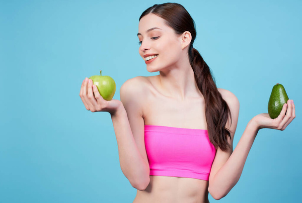 Attractive athletic young girl, in a sports pink top, holds an avocado and a green apple in her hands, smiles and enjoys looking at the apple in her right hand, stands on a blue background. - Фото, изображение