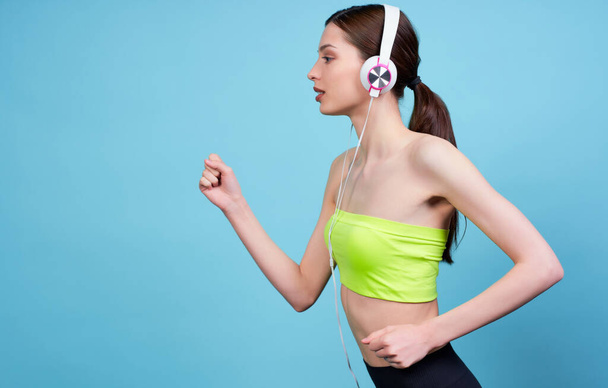 Portrait of a serious beautiful attractive woman athlete, in light green top and black leggings, hair is piled in tail, running with headphones to the left, isolated on blue background. Copy space. - Foto, imagen