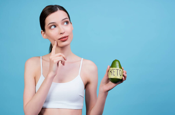 Pensive attractive pretty young sports girl in a white top, holding an avocado in her left hand, on which a tape with the word keto is glued, a girl on a keto diet, posing on a blue background. - Foto, Bild