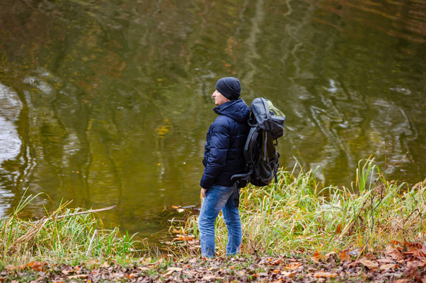 Traveller with backpack is standing on the bank of the river with azure water at sunny day in autumn. Travel. Landscape with fit man, orange trees in fall. Hiking concept. View from the back - Foto, imagen