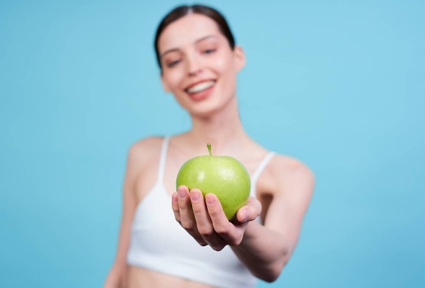 Cute attractive smiling young Caucasian girl, in a white top, holds a juicy green apple on her outstretched arm, posing on a blue background. The concept of a healthy diet, fruits. - Photo, image