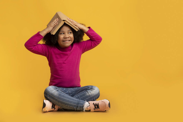 Piękny mały czarny dziecko siedzi na podłodze z książki na głowie - Zdjęcie, obraz