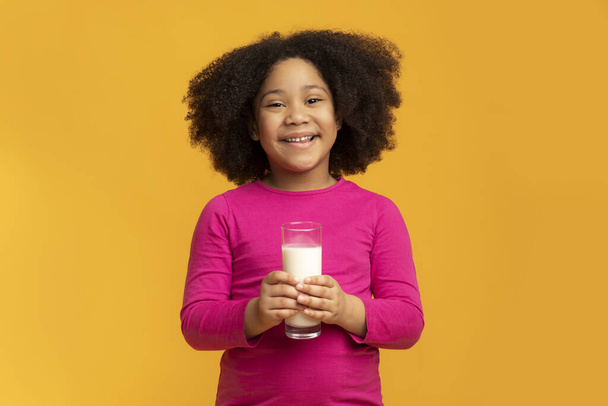 Cute afro girl holding glass of milk, standing on yellow background - Fotó, kép