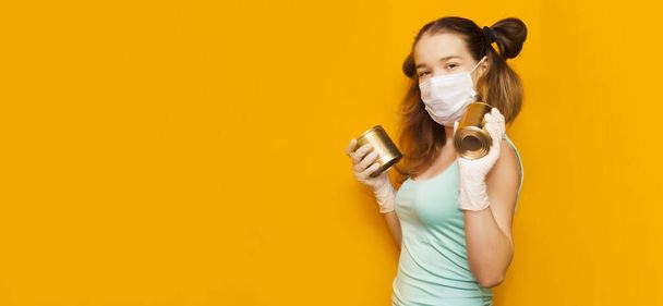 a young girl in a mask and gloves on an orange background with two cans of canned food in her hands. Funny and with ponytailsProtection AGAINST covid-19 coronavirus - Fotó, kép