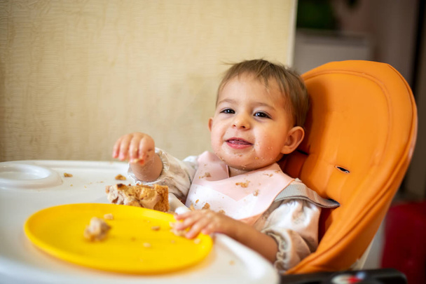 可愛い赤ちゃんは汚い手と顔のテーブルがあるオレンジのベビーチェアに座ってカメラを見て笑います。テーブルの上にはパン粉とオレンジのプレート。クローズアップ、サイドビュー、ソフトフォーカス、ぼかしの背景 - 写真・画像